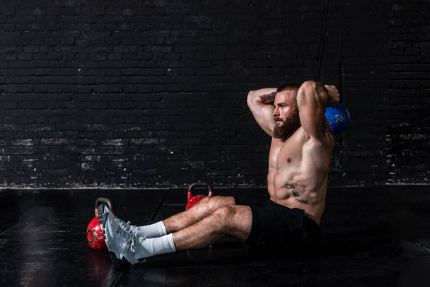 Young sweaty strong muscular fit man with big muscles sitting on the floor of the gym and doing triceps workout with the heavy kettlebell as hardcore cross training - 写真・画像