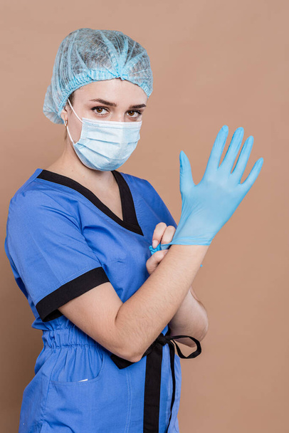 Молодой врач в хирургической форме с защитной фуражкой на голове и голубыми перчатками на фоне персика. - Фото, изображение