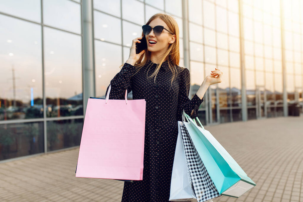 Happy young woman in sunglasses, with shopping bags, talking on a mobile phone in the background of a shopping center - Foto, Imagen