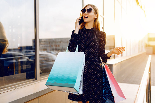 Happy beautiful young woman in sunglasses, with shopping bags, talking on a mobile phone in the background of a shopping center - Foto, Imagem