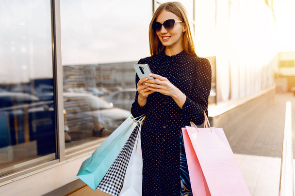 gelukkig mooi jong vrouw in zonnebril, met boodschappentassen, maakt gebruik van een mobiele telefoon, staan in de stad tegen de achtergrond van een winkelcentrum - Foto, afbeelding