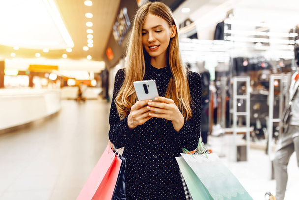 schöne junge Frau in einem Kleid, mit Einkaufstaschen, benutzt ein Mobiltelefon, während in einem Einkaufszentrum - Foto, Bild