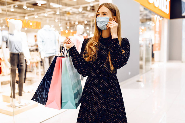 happy young woman in a medical protective mask on her face, with shopping bags, walks through the shopping center - Photo, image