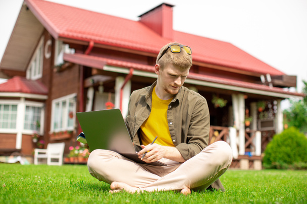 Босоногий юный студент сидит на зеленой траве с ноутбуком обдумывает текст - Фото, изображение