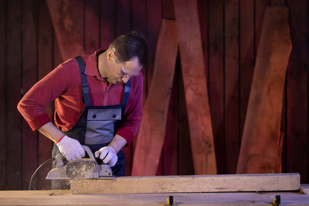 плотник среднего возраста обрабатывает деревянную деталь с помощью электрического агрегата - Фото, изображение