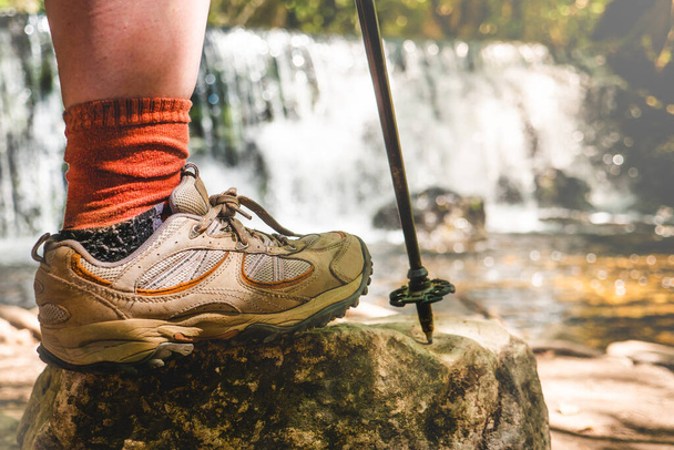 Donna gamba con scarponi da trekking e bastone su una roccia con una cascata naturale dietro.  - Foto, immagini