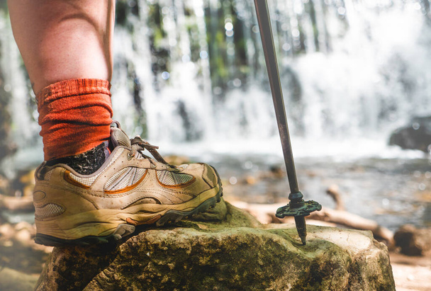 Pierna de mujer con botas de senderismo y palo en una roca con una cascada natural detrás.  - Foto, Imagen