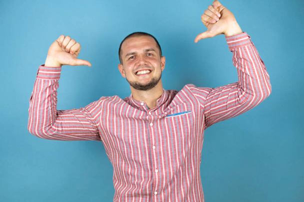 Russischer Geschäftsmann, der rotes Hemd über blauem Hintergrund trägt, sieht selbstbewusst mit einem Lächeln im Gesicht aus und zeigt stolz und glücklich mit den Fingern. - Foto, Bild