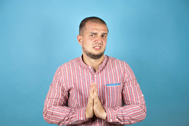 Homme d'affaires russe portant chemise rouge sur fond bleu mendier et prier avec les mains ainsi que l'expression de l'espoir sur le visage très émotionnel et inquiet - Photo, image
