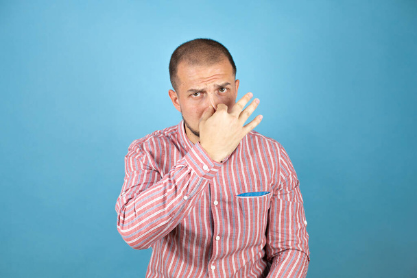 Hombre de negocios ruso con camisa roja sobre fondo azul oliendo algo apestoso y asqueroso, olor intolerable, conteniendo la respiración con los dedos en la nariz - Foto, imagen