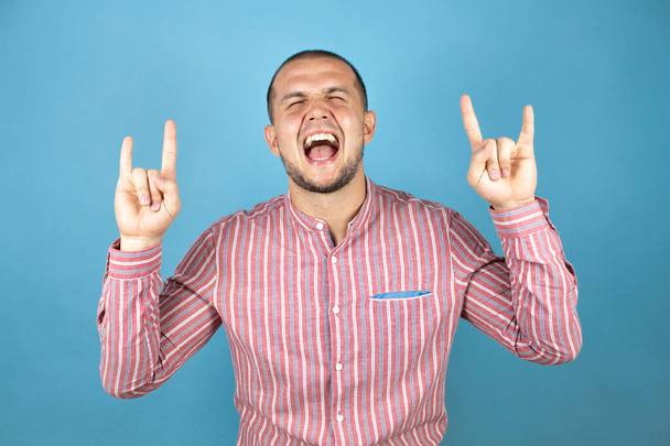 Hombre de negocios ruso sobre fondo azul gritando con expresión loca haciendo símbolo de roca con las manos arriba. Estrella de la música. Concepto pesado. - Foto, Imagen