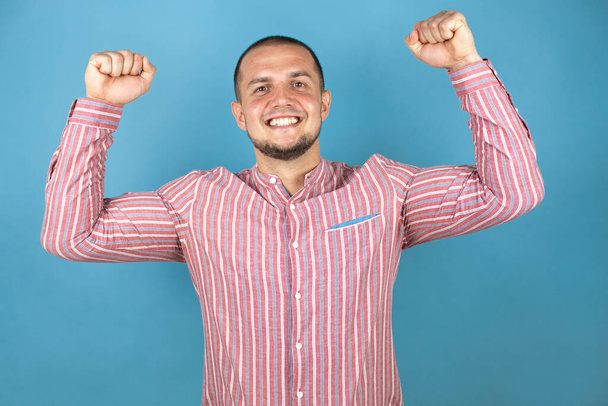 Ruský obchodník nad modrým pozadím velmi šťastný a vzrušený dělá vítězný gesto se zdviženýma rukama, usmívá se a křičí po úspěchu. Koncept oslavy. - Fotografie, Obrázek