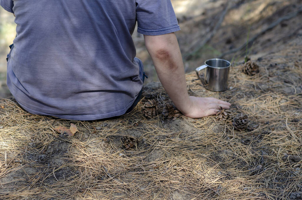 Muž sedí na zemi v lese. Kovový hrnek s pitím poblíž. Pohled zezadu. Pěší turistika. Aktivní životní styl. - Fotografie, Obrázek