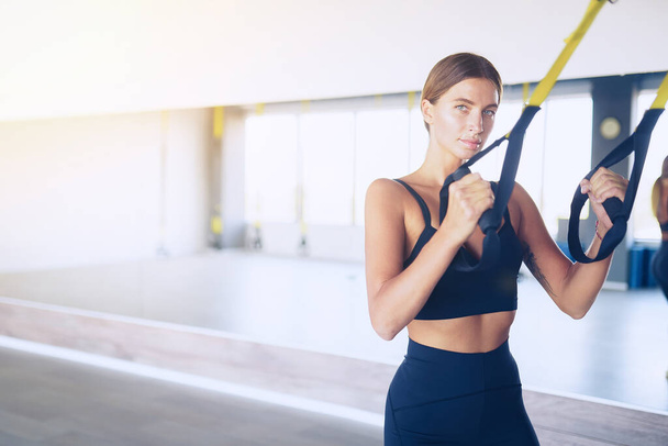 Beautiful young woman training with suspension trainer sling or suspension straps in gym. Upper body exercise concept on TRX - Foto, Imagem