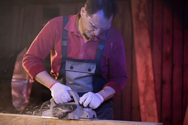 40s samec tesař zpracovává dřevěný obrobek s elektrickým hoblíkem - Fotografie, Obrázek