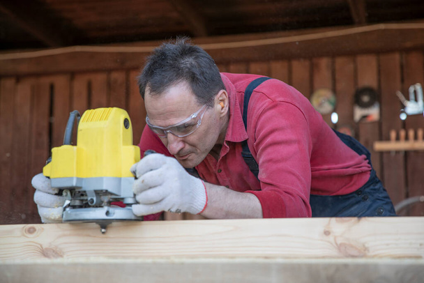 Красивый плотник средних лет работает на деревянной детали с помощью инструмента фрезерования - Фото, изображение