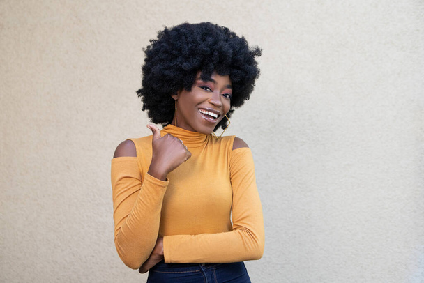 Photo of African American woman with curly hair and happy face, showing thumb up, wearing casual wear, expresses positive emotions, feels entertained, posing on light background with empty copy space. - Фото, зображення