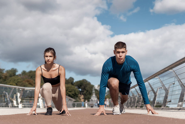 Focused young adult woman and serious man ready for competition. Friends making sport training outdoors together, preparing for running, sitting near start finish line - Photo, Image