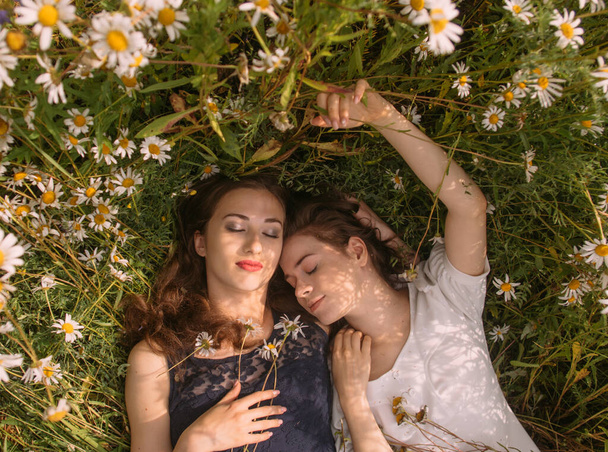 Deux filles avec les yeux fermés dans des robes bleu foncé et blanc dans la journée ensoleillée couché dans le champ de camomille sur la nature - Photo, image