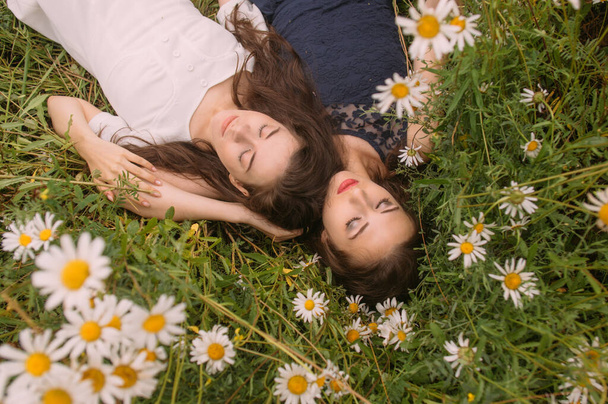 Dos chicas con los ojos cerrados en azul oscuro y vestidos blancos en el día soleado acostado y durmiendo en el campo de manzanilla en la naturaleza - Foto, imagen