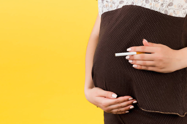 Sarı arka planda göbeğine sigara dayamış kahverengi pijamalı hamile kadını yakın plan. Doğmamış bebek için tehlikeli bir risk. - Fotoğraf, Görsel