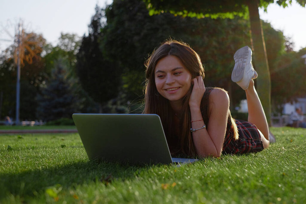 Young woman using laptop in the park lying on the green grass. Leisure time activity concept.  - 写真・画像