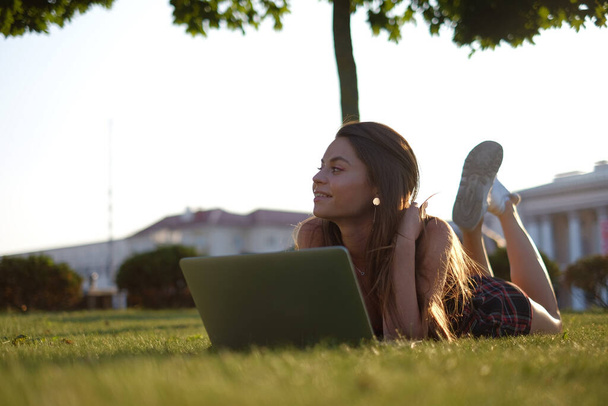 Young woman using laptop in the park lying on the green grass. Leisure time activity concept.  - Фото, изображение