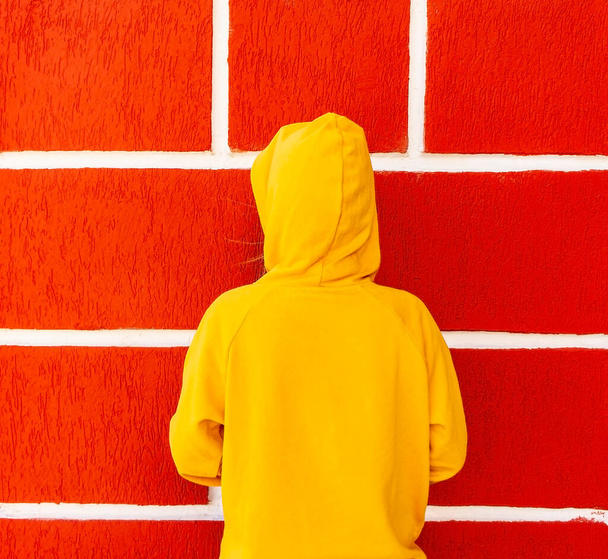tiener meisje in een gele sweater of hoodie in een kap tegen de achtergrond van een rode muur met witte naden - Foto, afbeelding