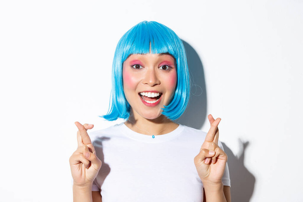 Close-up of hopeful cute asian girl in blue wig making wish, cross fingers good luck, standing over white background - Photo, Image