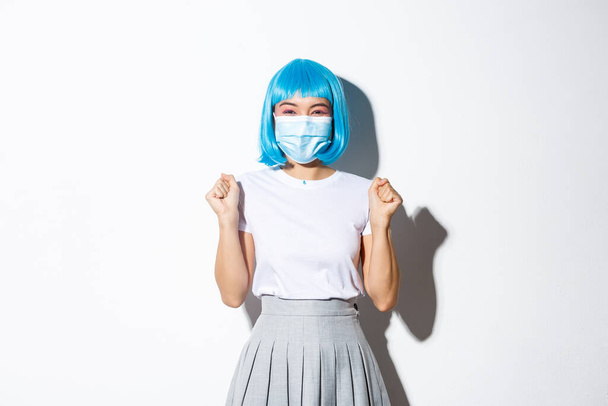 Concept of halloween celebration and covid-19 pandemic. Happy asian girl enjoying party, wearing blue wig and medical mask, jumping from happiness, triumphing over white background - Foto, immagini