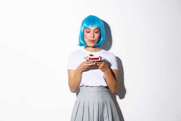 Imagen de chica asiática en peluca azul lamiendo labios de la tentación, mirando el pedazo de pastel, de pie sobre fondo blanco - Foto, Imagen
