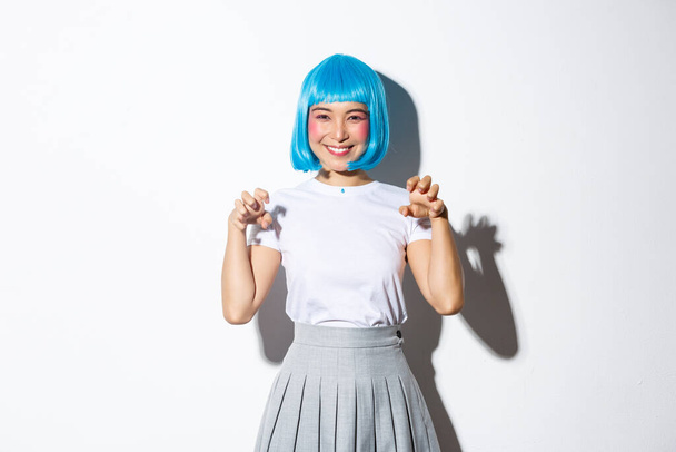 Hauska ja söpö aasialainen tyttö sinisessä peruukissa hymyilee ja tekee käsi tassut ele, juhlii halloween, seisoo valkoisella taustalla - Valokuva, kuva