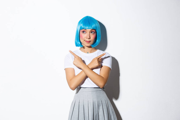 Image of pretty asian girl in blue wig pointing fingers sideways, looking curious while making choice, standing in halloween costume over white background - Photo, Image