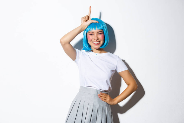 Portrait of beautiful silly asian girl celebrating halloween, showing loser gesture on forehead and smiling, mocking someone, standing over white background - Photo, Image