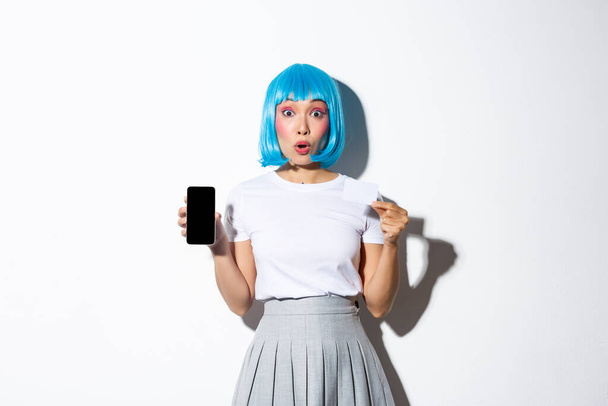 Πορτρέτο του έκπληκτος κορίτσι αίγλη σε μπλε περούκα, λαχανιάσει κατάπληκτος, δείχνει πιστωτική κάρτα και οθόνη smartphone, στέκεται πάνω από το λευκό φόντο - Φωτογραφία, εικόνα