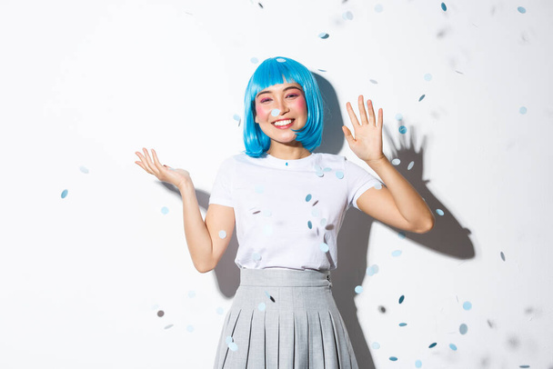 Cheerful asian girl in blue wig throwing confetti over white background, celebrating halloween, waving hand to say hello, greeting someone - Foto, Bild
