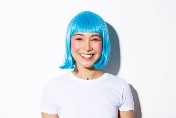 Close-up of cute asian female celebrating halloween in blue wig, smiling happy at camera, standing over white background - Foto, imagen