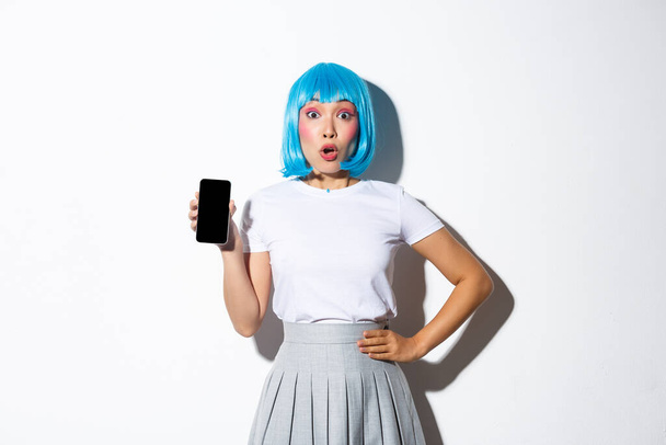 Portret van verbaasd mooi meisje in blauwe pruik toont iets cool op smartphone scherm, staande over witte achtergrond - Foto, afbeelding