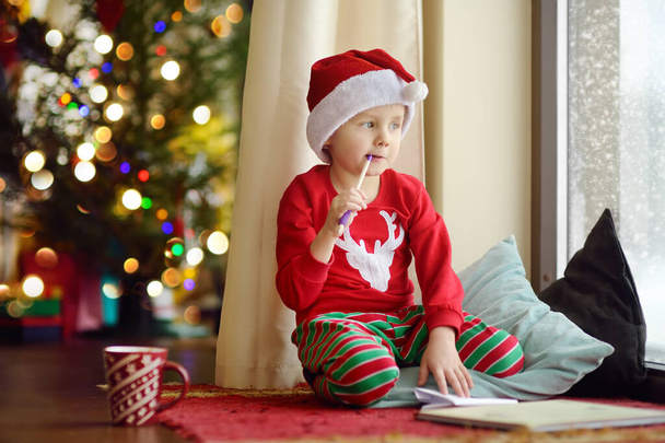 Little boy writing the letter to Santa and look out the window at the snowfall. Child dreams of a gift that he can receive. Magic mail of Santa Claus. Traditions around New Year and Christmas Holidays - Photo, image
