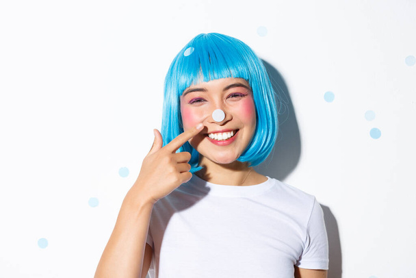 Image of cute and silly asian girl with confetti on her nose smiling and looking happy, wearing blue wig for halloween party - Photo, Image