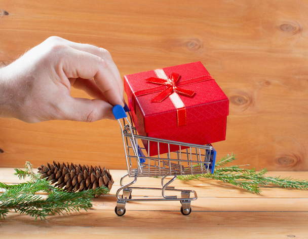 men hand keep miniature shopping cart on wheels loaded with boxes of gifts. wooden background with branches of a Christmas tree and cones. concept gifts for christmas and new year. copy space. - Photo, Image