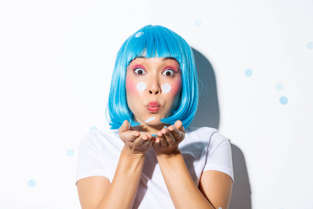 Close-up van aantrekkelijke aziatische meisje in blauwe pruik blazen confetti, vieren Halloween, staande over witte achtergrond - Foto, afbeelding