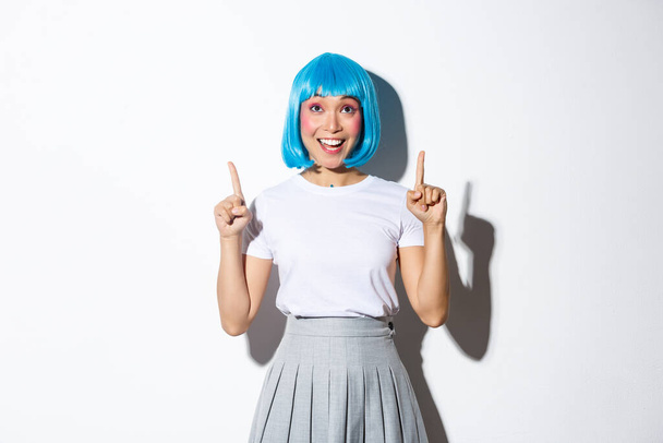 Muotokuva onnellinen hymyilevä aasialainen tyttö sininen puolue peruukki näköinen, osoittaa sormella ylös, näyttää logon halloween tai loma juhla, seisoo valkoisella taustalla - Valokuva, kuva