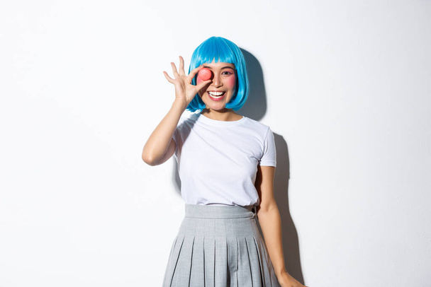 Cute asian girl in anime wig and schoolgirl uniform looking happy, showing macaron and smiling, standing over white background - Fotografie, Obrázek