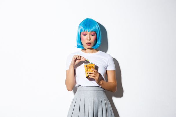 Retrato de chica asiática coqueta en peluca azul celebrando halloween, comiendo dulces de truco o bolsa de regalo, de pie sobre fondo blanco - Foto, Imagen