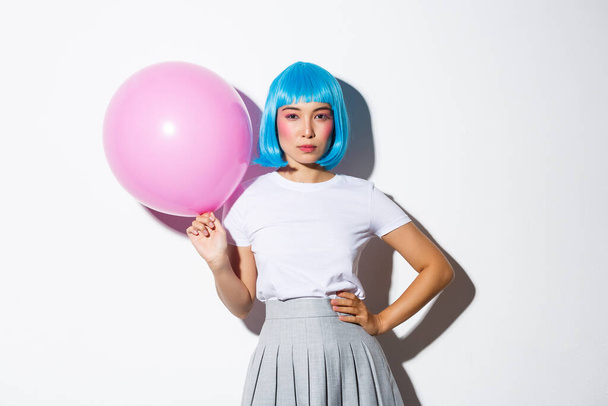 Image of sassy asian woman in blue wig, looking determined, holding pink balloon, standing over white background - Фото, изображение