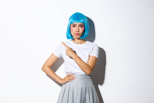 Image of skeptical and unamused asian girl in blue party wig, grimacing upset and pointing finger at upper left corner, standing against white background - Foto, afbeelding