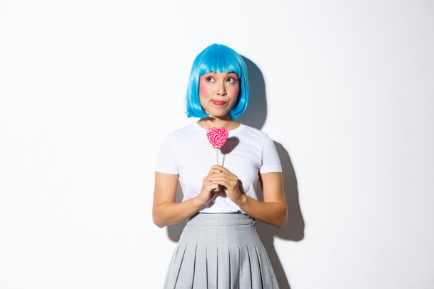 Imagen de chica asiática tonta en peluca azul, vestida para la fiesta de halloween, luciendo soñadora en la esquina superior izquierda, sosteniendo dulces en forma de corazón - Foto, Imagen