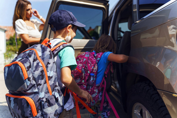 Little boy and girl in sunny day or morning going to school - small pupils first graders with backpack on shoulders and their mother entering the car - Real people education concept - Foto, Imagen