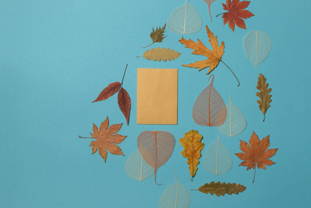 Μπλε φόντο με ξηρά φθινοπωρινά φύλλα και μια χάρτινη κάρτα στο κέντρο, με χώρο για κείμενο - Φωτογραφία, εικόνα
